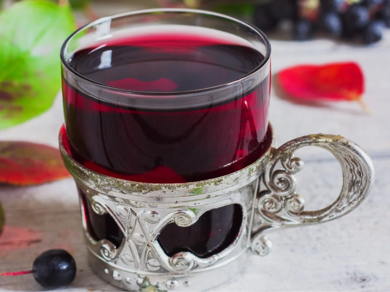 Aronia ma wiele witamin i wartości. Doskonale smakuje w formie naparu. Szklanka z ciemnoczerwonym naparem z aronii w metalowym koszyczku, obok liście i owoce aronii.