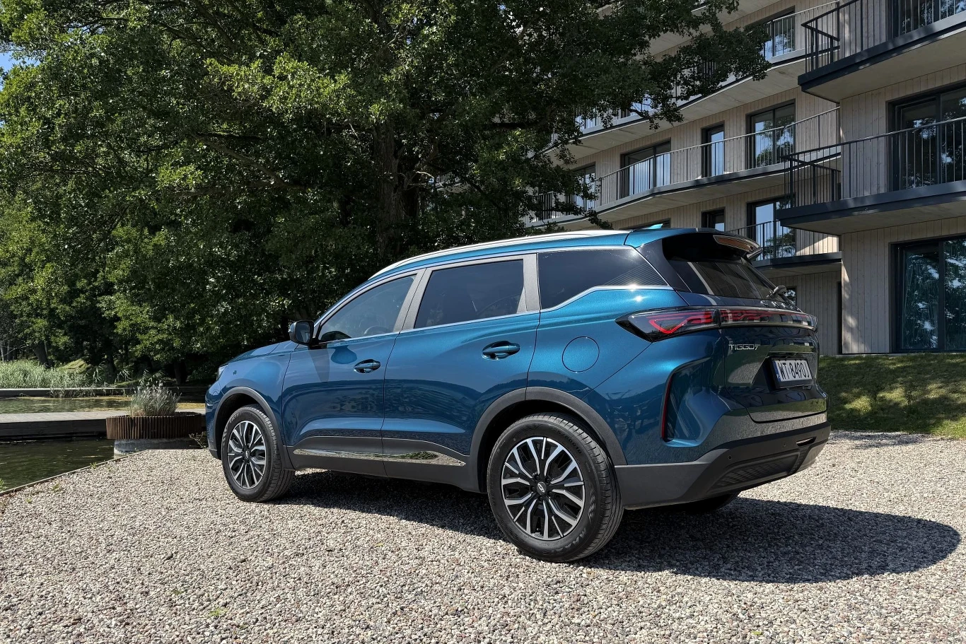 Niebieski SUV z przyciemnianymi szybami i nowoczesnym designem zaparkowany na żwirowym placu obok budynku z balkonami, w otoczeniu zieleni.