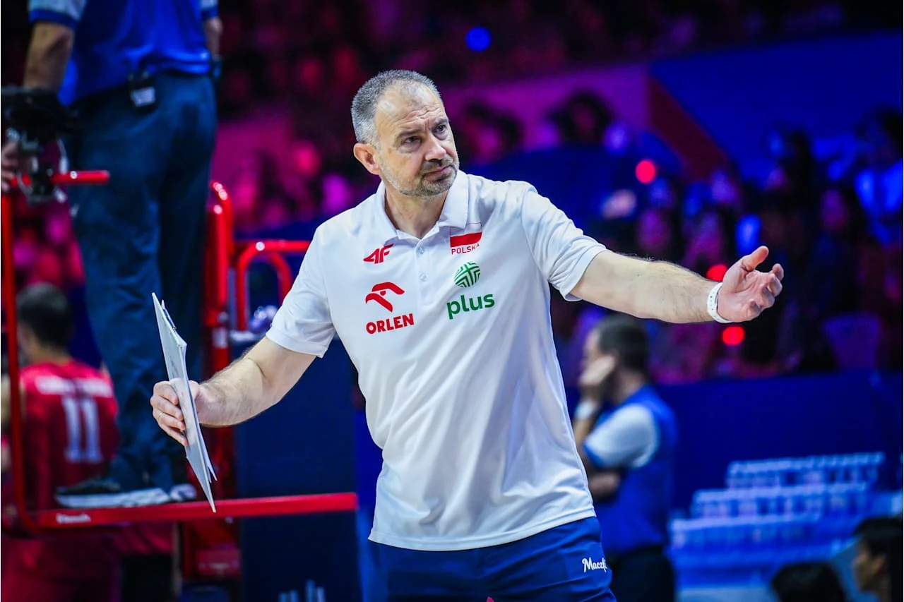 Nikola Grbić Trener polskiej reprezentacji siatkarzy w białej koszulce z logotypami sponsorów gestykuluje podczas meczu, trzymając kartki w lewej ręce.