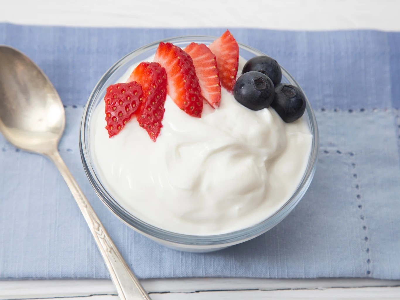 Jogurt naturalny czy grecki? Który z nich jest lepszy? Szklana miseczka z gęstym jogurtem naturalnym, plasterkami truskawek i jagodami, obok srebrna łyżka.