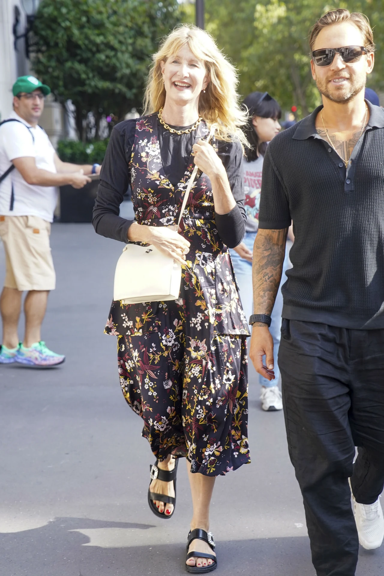 Laura Dern w Paryżu. Aktorka zachwyciła w kwiecistej sukience Uśmiechnięta kobieta w długiej, kwiecistej sukience i mężczyzna w czarnej koszuli idą razem ulicą; kobieta niesie jasną torebkę na ramię, a mężczyzna ma okulary przeciwsłoneczne.