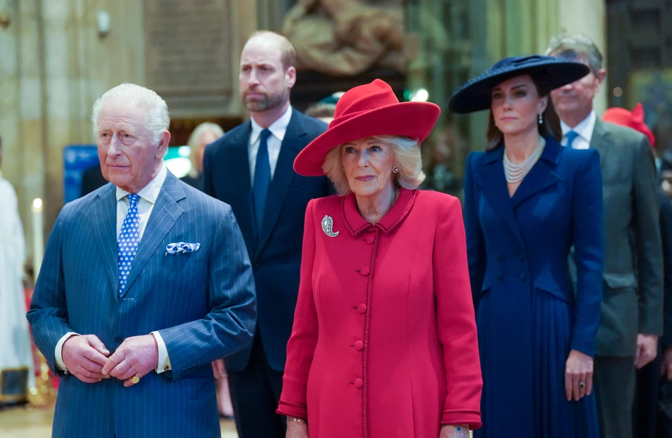 Cztery elegancko ubrane osoby stoją w rzędzie w przestronnym, reprezentacyjnym wnętrzu; mężczyzna z siwymi włosami w niebieskim garniturze, kobieta w czerwonym płaszczu i kapeluszu, mężczyzna w garniturze oraz kobieta w granatowej sukni i szerokim kape...