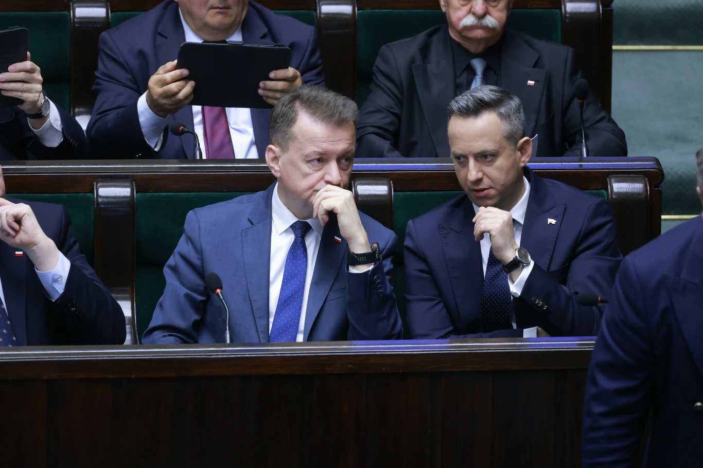 Dwóch mężczyzn w garniturach siedzi obok siebie na ławach sejmowych, pogrążonych w poważnej rozmowie, z dłońmi przy twarzach, w tle inni uczestnicy obrad.
