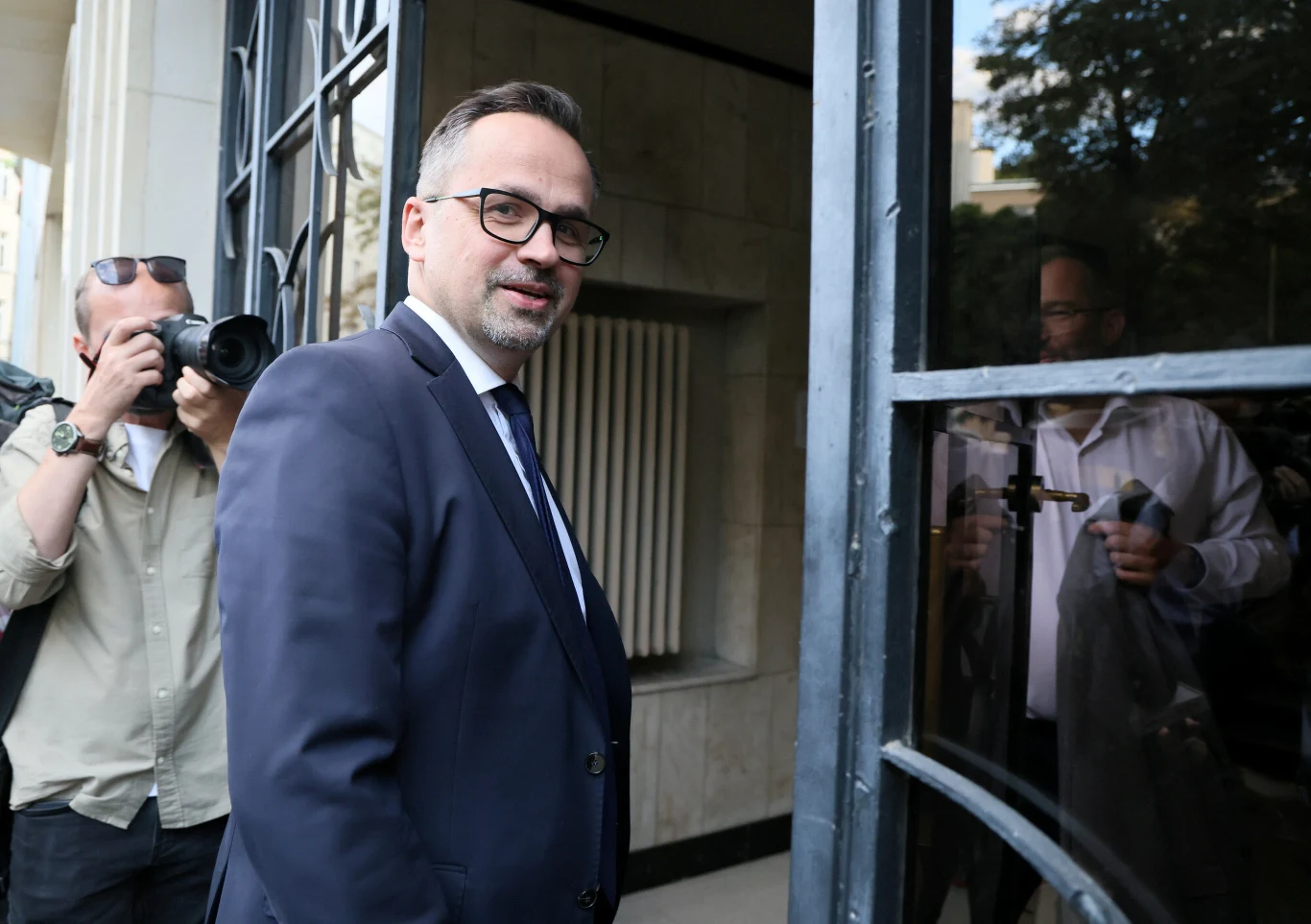 Mężczyzna w garniturze otwiera szklane drzwi, obok stoi fotograf wykonujący zdjęcie.