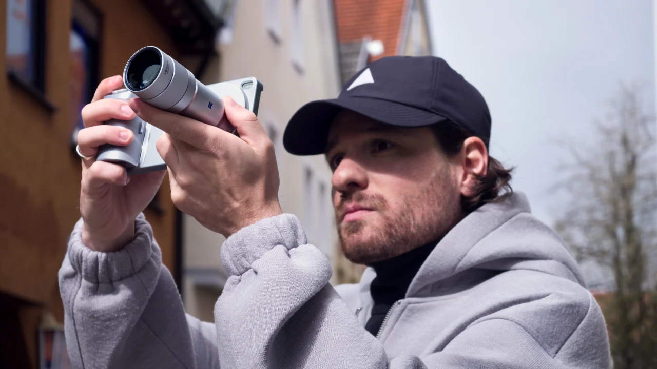 Smartfony z serii vivo X300 łączą poręczną konstrukcję z zaawansowanymi możliwościami fotograficznymi wspieranymi przez AI Mężczyzna w szarej bluzie i czapce z daszkiem fotografuje ulicę nowoczesnym aparatem trzymanym przy oku.