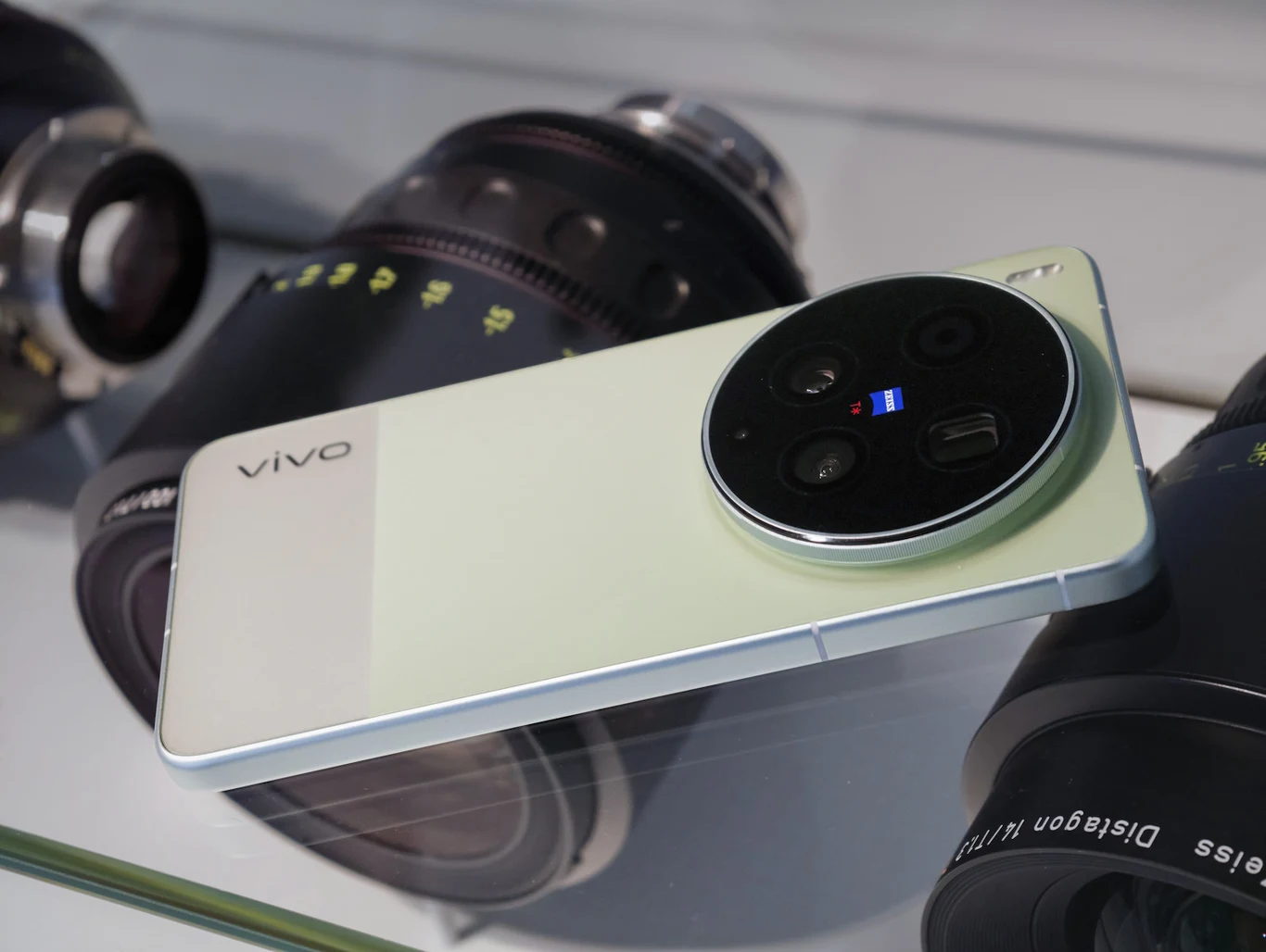 Smartfon vivo X300 Ultra to model dla twórców, którzy chcą mieć profesjonalne możliwości fotograficzne i filmowe zawsze w kieszeni Smartfon Vivo z dużą, okrągłą wyspą aparatów i logo Zeiss leży na przezroczystej powierzchni obok kilku profesjonalnych obiektywów fotograficznych.