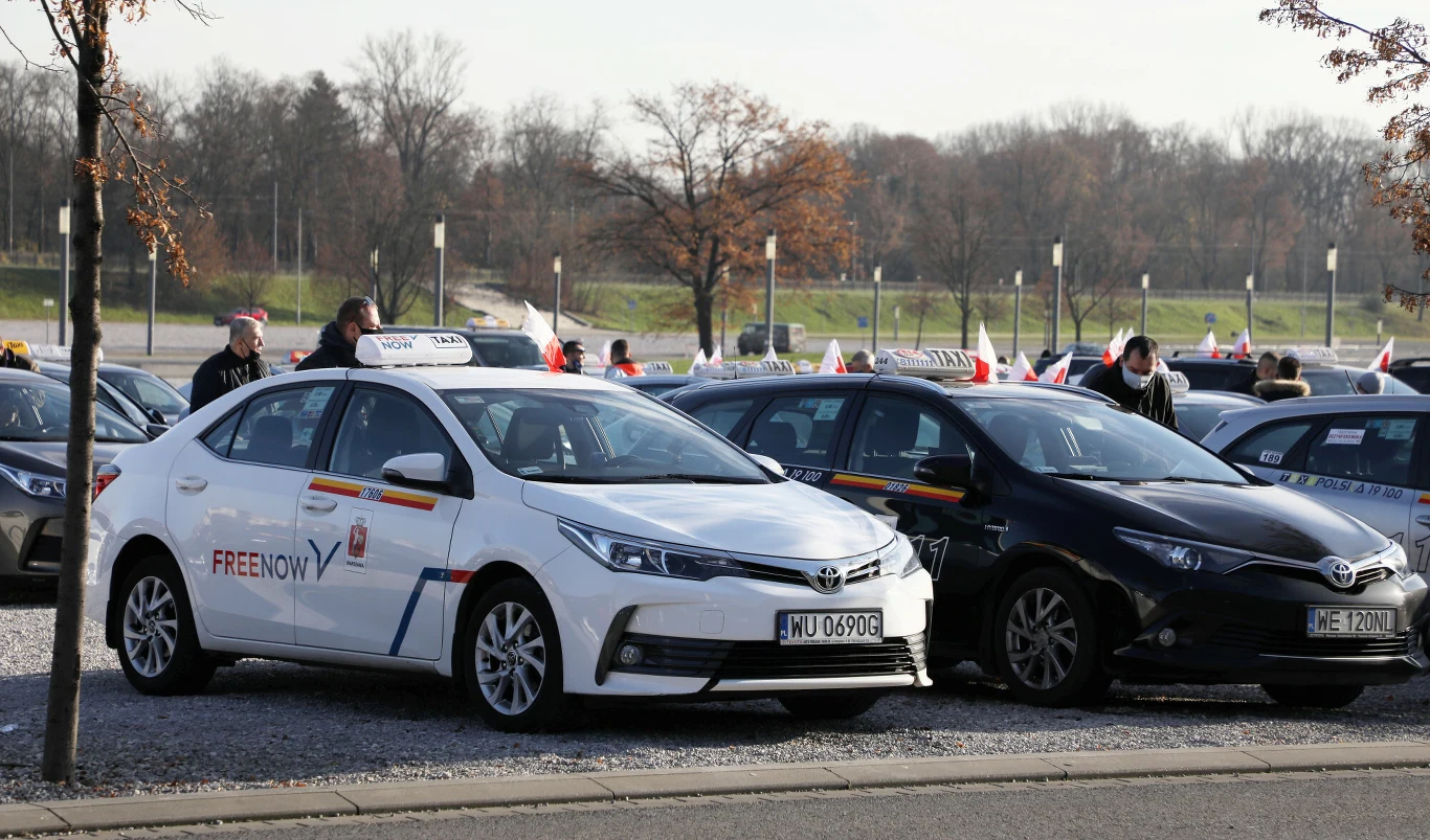 Szereg zaparkowanych samochodów osobowych z oznaczeniami taxi oraz widocznymi biało-czerwonymi flagami, przy których stoją osoby rozmawiające lub przygotowujące pojazdy.