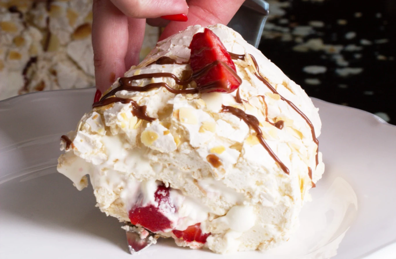 Rolada bezowa z truskawkami i kremem mascarpone smakuje jak kwintesencja lata Rolada bezowa z kremem mascarpone, świeżymi truskawkami i polewą czekoladową, trzymana przez osobę z czerwonymi paznokciami.