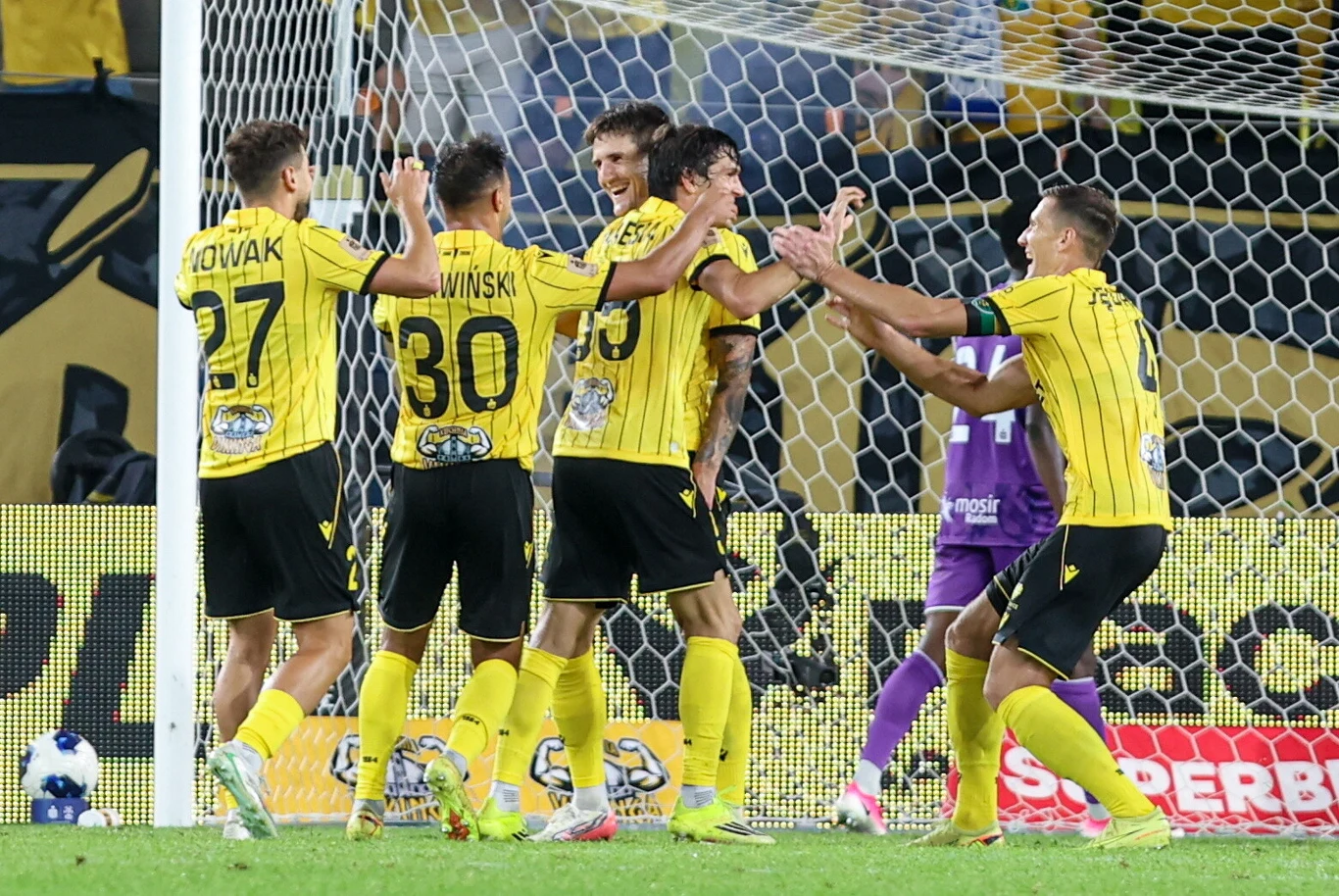 Grupa piłkarzy w żółto-czarnych strojach świętuje zdobycie gola przy bramce, otoczona przez radośnie reagujących kolegów z zespołu.