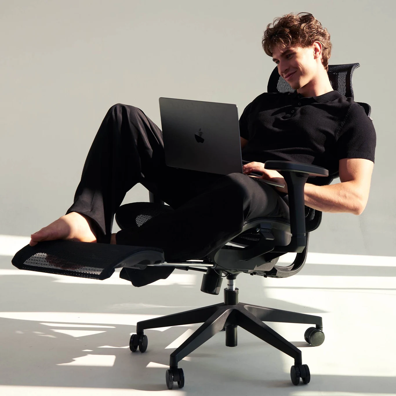 Młody mężczyzna w czarnym ubraniu relaksuje się na ergonomicznym fotelu biurowym, z nogami wyciągniętymi na podnóżku, pracując na laptopie.