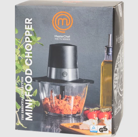 Opakowanie rozdrabniacza do żywności MasterChef z wyeksponowanym sprzętem wypełnionym posiekanymi składnikami, w otoczeniu oliwy z oliwek, pomidorków koktajlowych, ziół i deski z czosnkiem.