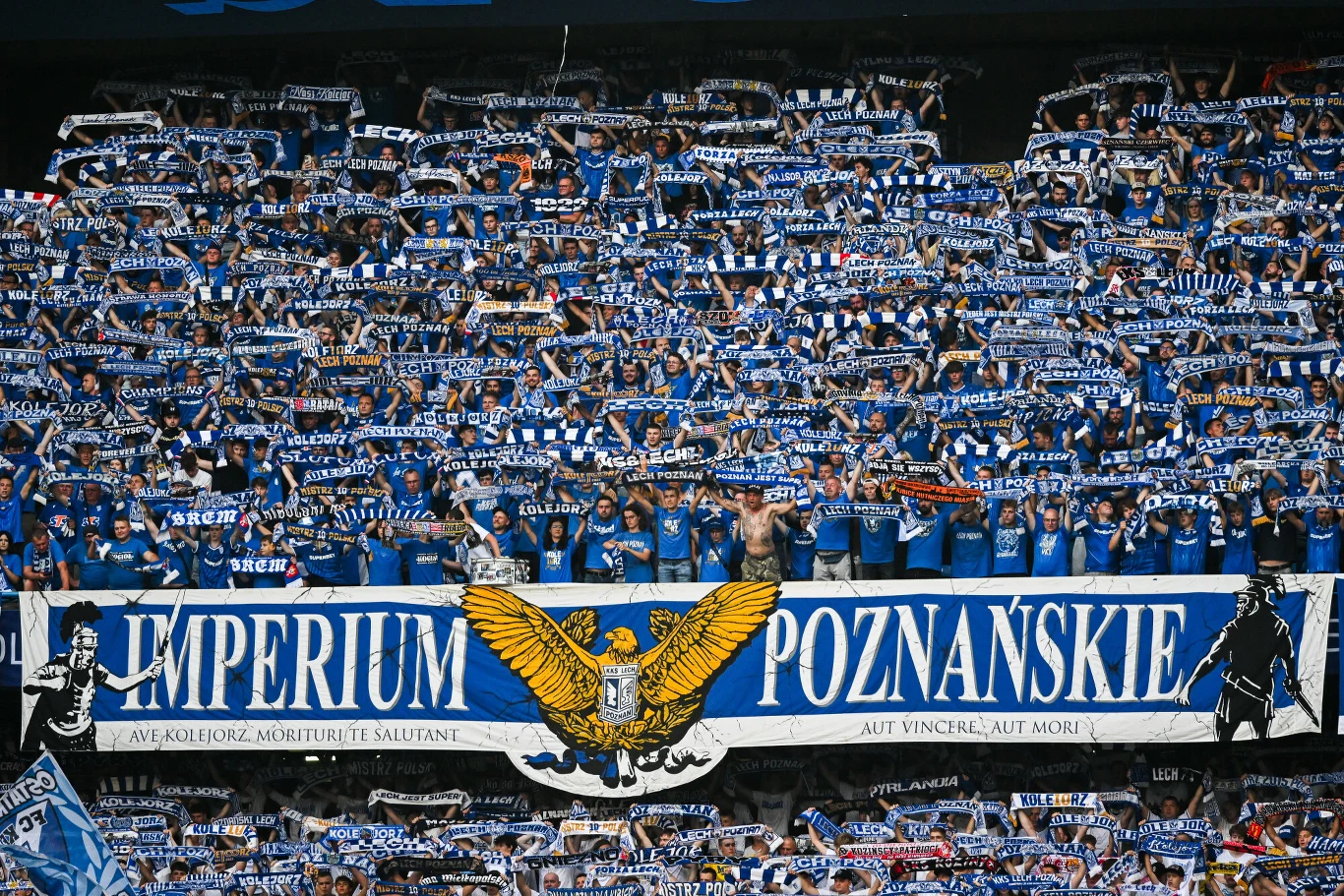 Kibice Lecha Poznań Tłum ubranych na niebiesko kibiców podnoszących klubowe szaliki w górę oraz duży transparent z napisem „IMPERIUM POZNAŃSKIE” i wizerunkiem orła pośrodku.