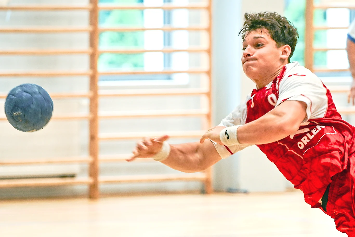 Zawodnik w czerwonej koszulce rzuca piłkę ręczną podczas dynamicznej akcji na hali sportowej.