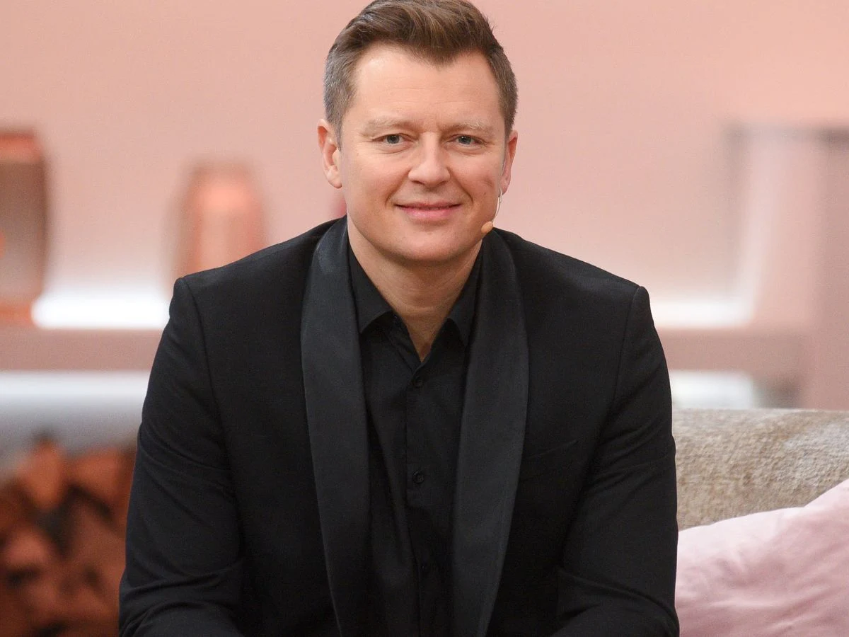 Rafał Brzozowski Rafał Brzozowski w czarnym garniturze siedzący na jasnej kanapie w nowoczesnym wnętrzu.