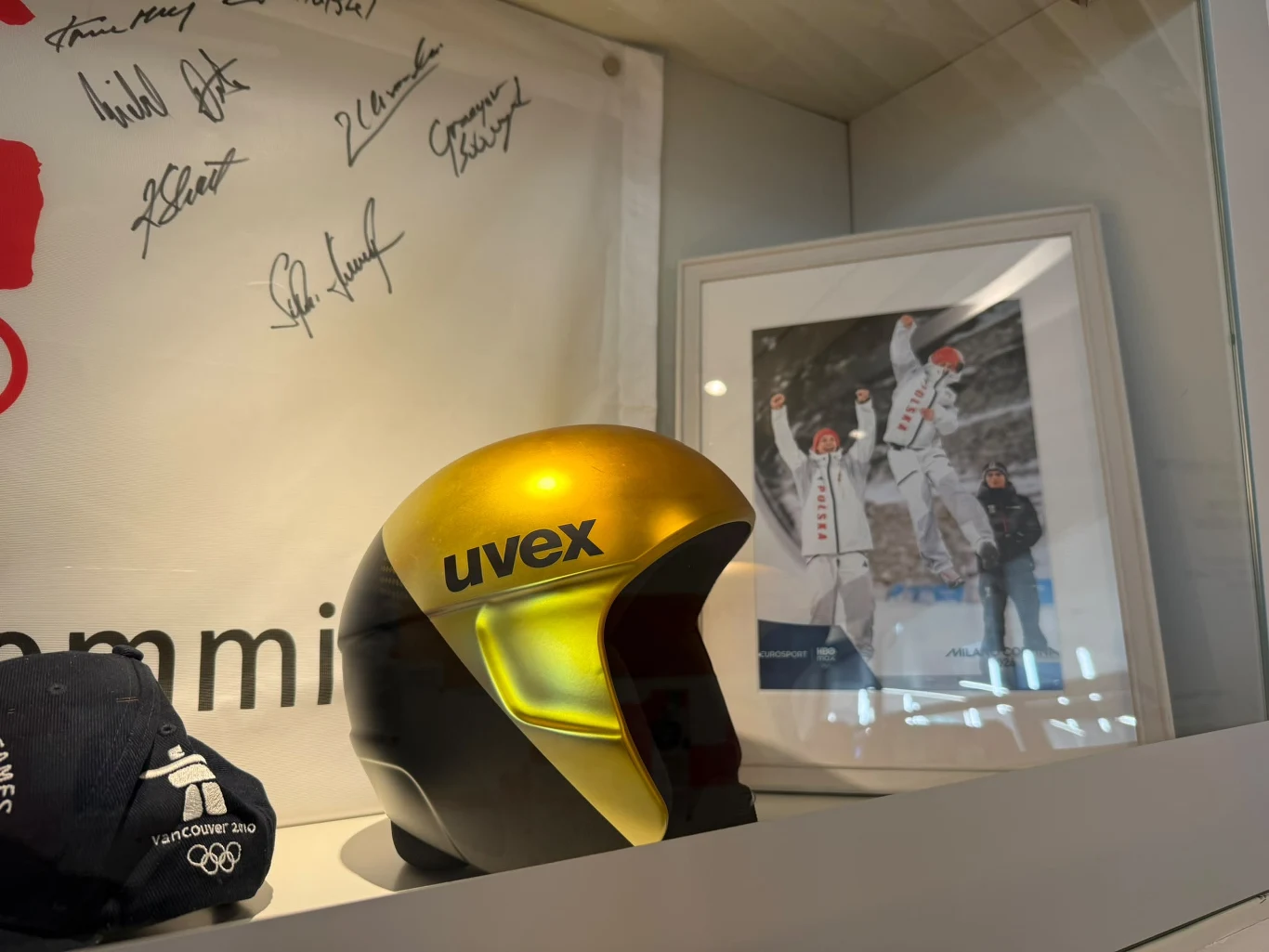Beskidzkie Centrum Narciarstwa Złoty kask marki Uvex ustawiony na półce obok pamiątkowej czapki olimpijskiej oraz oprawionego zdjęcia z podium olimpijskiego, w tle widoczny fragment białego materiału z autografami.