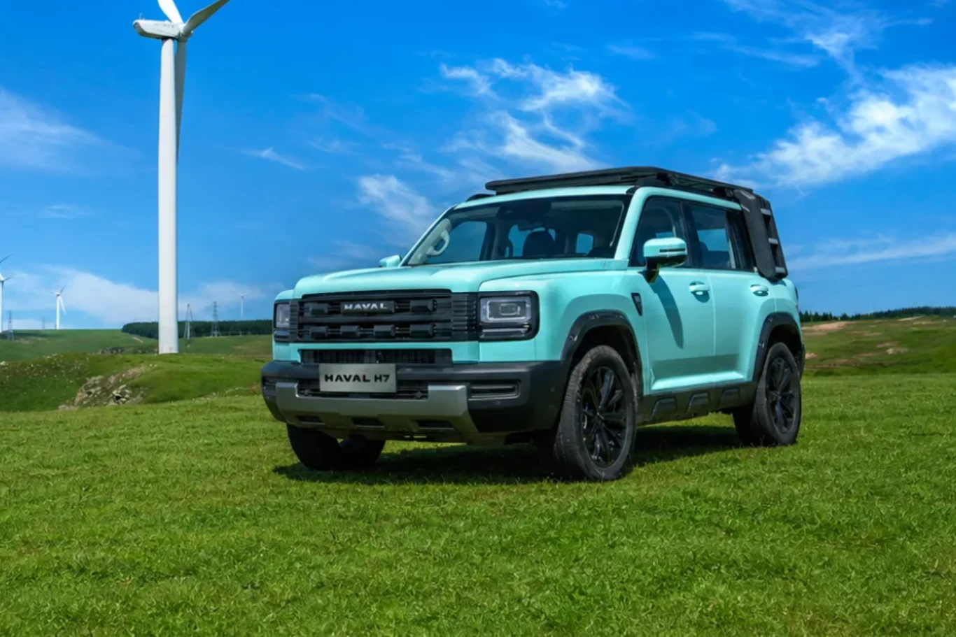 Kanciaste kształty Havala H7 mogą się podobać. Jasnoniebieski SUV Haval H7 stoi na zielonej trawie, w tle znajdują się nowoczesne turbiny wiatrowe na tle błękitnego nieba z delikatnymi chmurami.