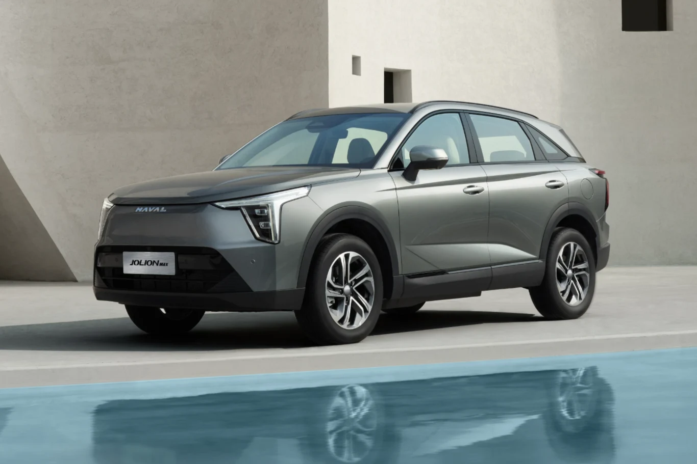 GWM Jolion MAX to rodzinny SUV o bezpiecznym, klasycznym wyglądzie. Srebrny SUV nowoczesnej linii stoi zaparkowany przy minimalistycznym budynku, odbijając się w tafli wody położonej na pierwszym planie.