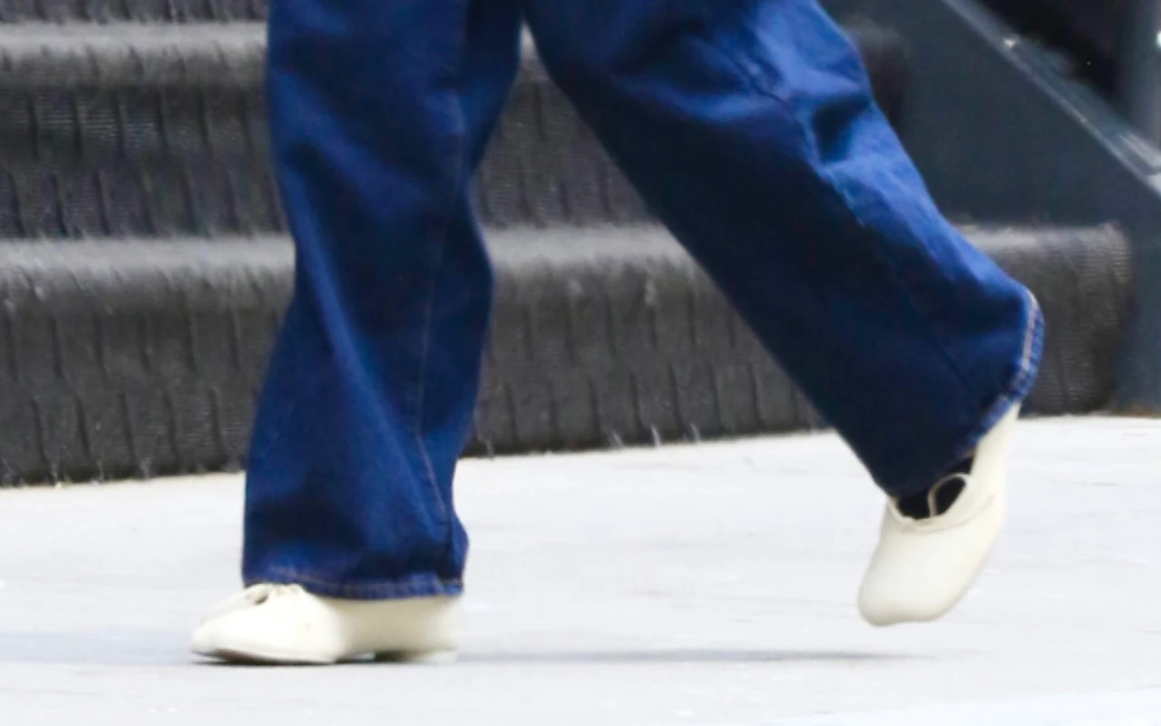 Białe sneakersy na stopach osoby ubranej w szerokie, niebieskie spodnie dżinsowe idącej po szarym chodniku obok schodów.