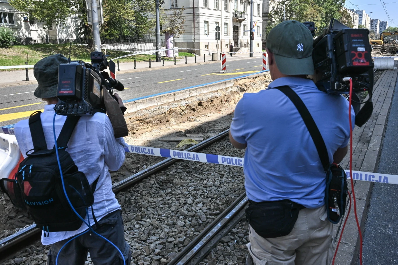 Dwóch operatorów kamer stojących przy torach tramwajowych, za policyjną taśmą zabezpieczającą wykop w miejscu prowadzenia prac służb.