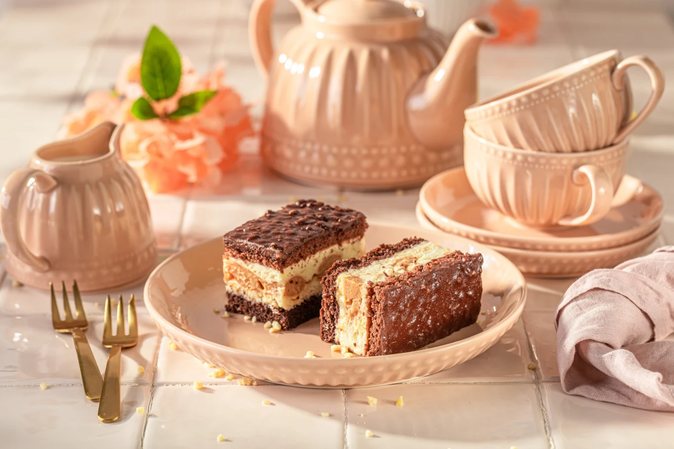 Ciasto Jeżyk smakuje jak popularne ciasteczka z karmelem, ryżem preparowanym i orzechami. Dwa kawałki ciasta Jeżyk z kremem na beżowym talerzyku, obok porcelanowy serwis do herbaty i złote widelczyki