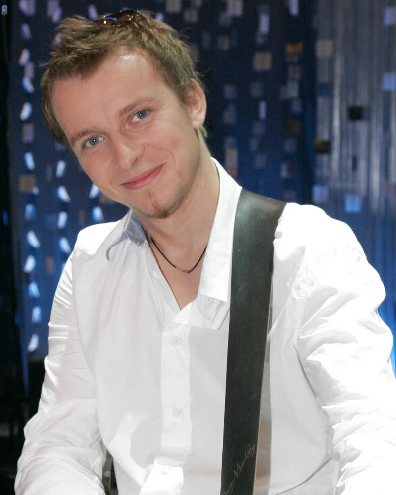 Piotr Kupicha w 2007 roku, u progu kariery Uśmiechnięty mężczyzna w białej koszuli z gitarą na ramieniu, na tle ciemnoniebieskich dekoracji.