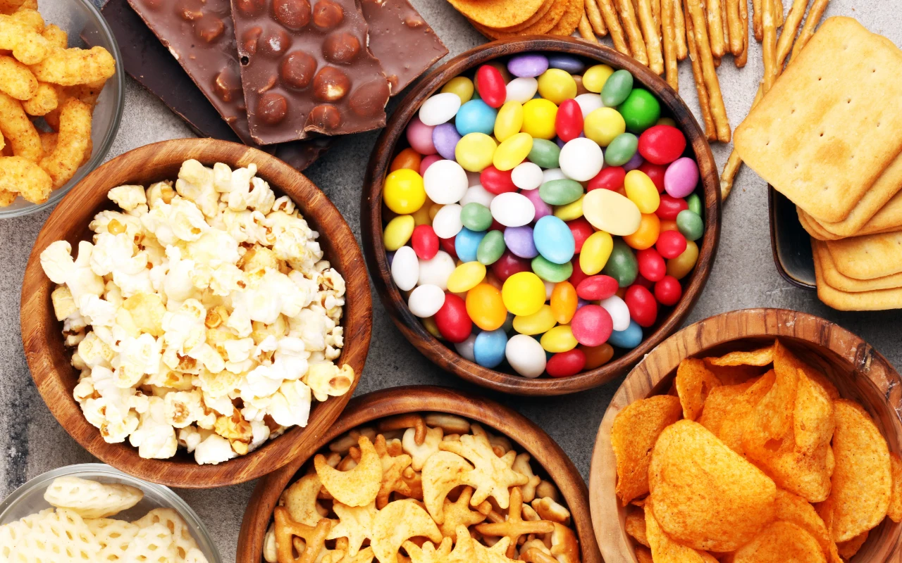 Miska kolorowych cukierków, miska popcornu, miska chipsów, kawałki czekolady, słone paluszki, krakersy oraz przekąski o różnych kształtach.