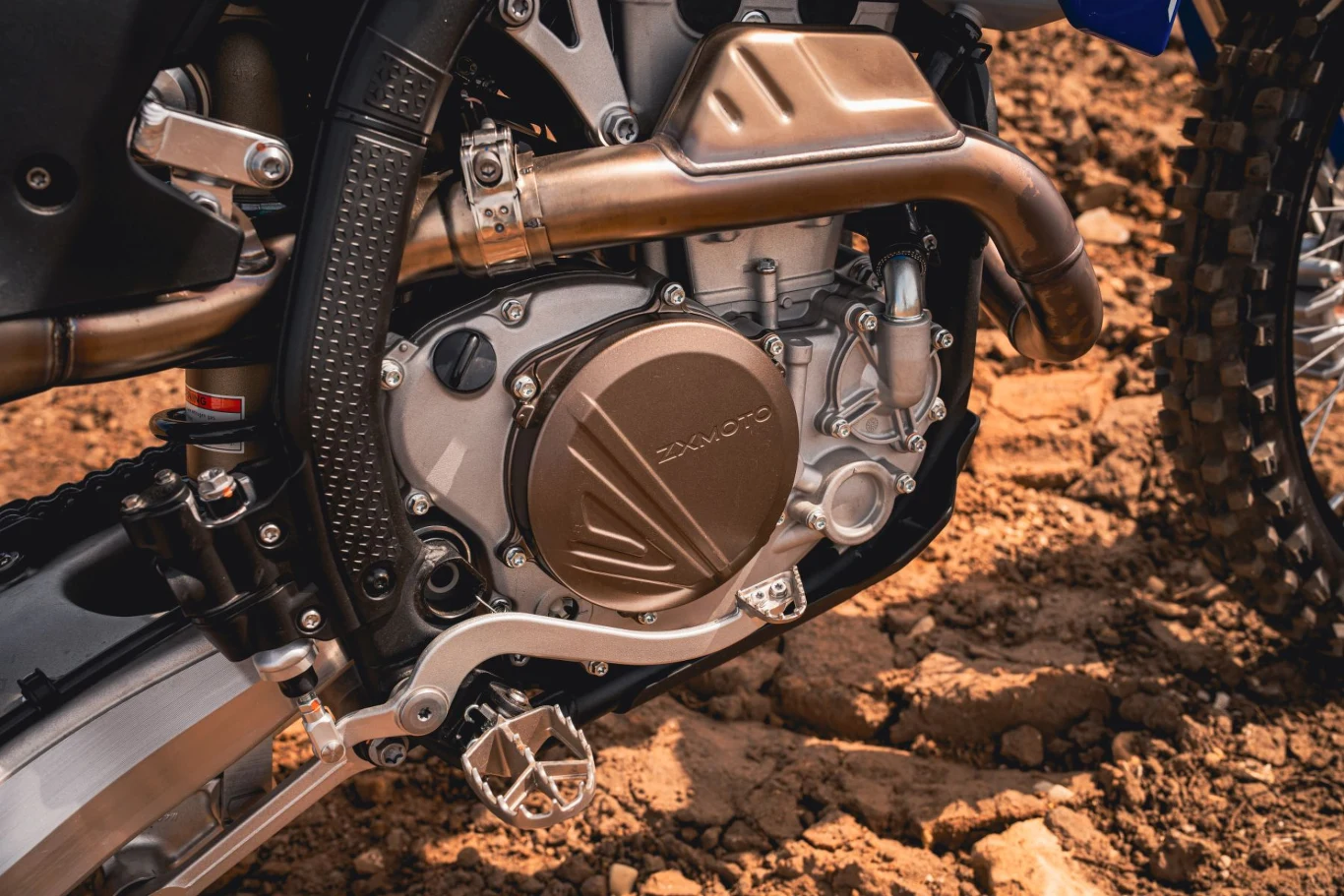 Detalicznie widoczny silnik motocykla terenowego z metalicznymi elementami konstrukcji, otoczony oponą z wyraźnym bieżnikiem i fragmentem ramy motocykla na tle ziemistego podłoża.