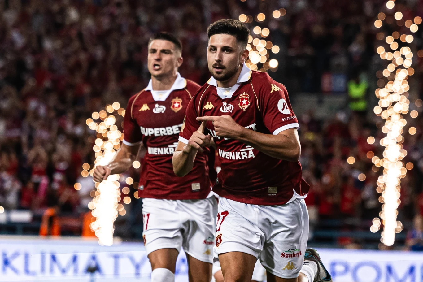 Jordi Sanchez i Marko Bozić w barwach Wisły Kraków podczas meczu PKO BP Ekstraklasy Jordi Sanchez i Marko Bozić z Wisły Kraków w bordowych koszulkach i białych spodenkach podczas meczu Ekstraklasy na tle fajerwerków i kibiców.