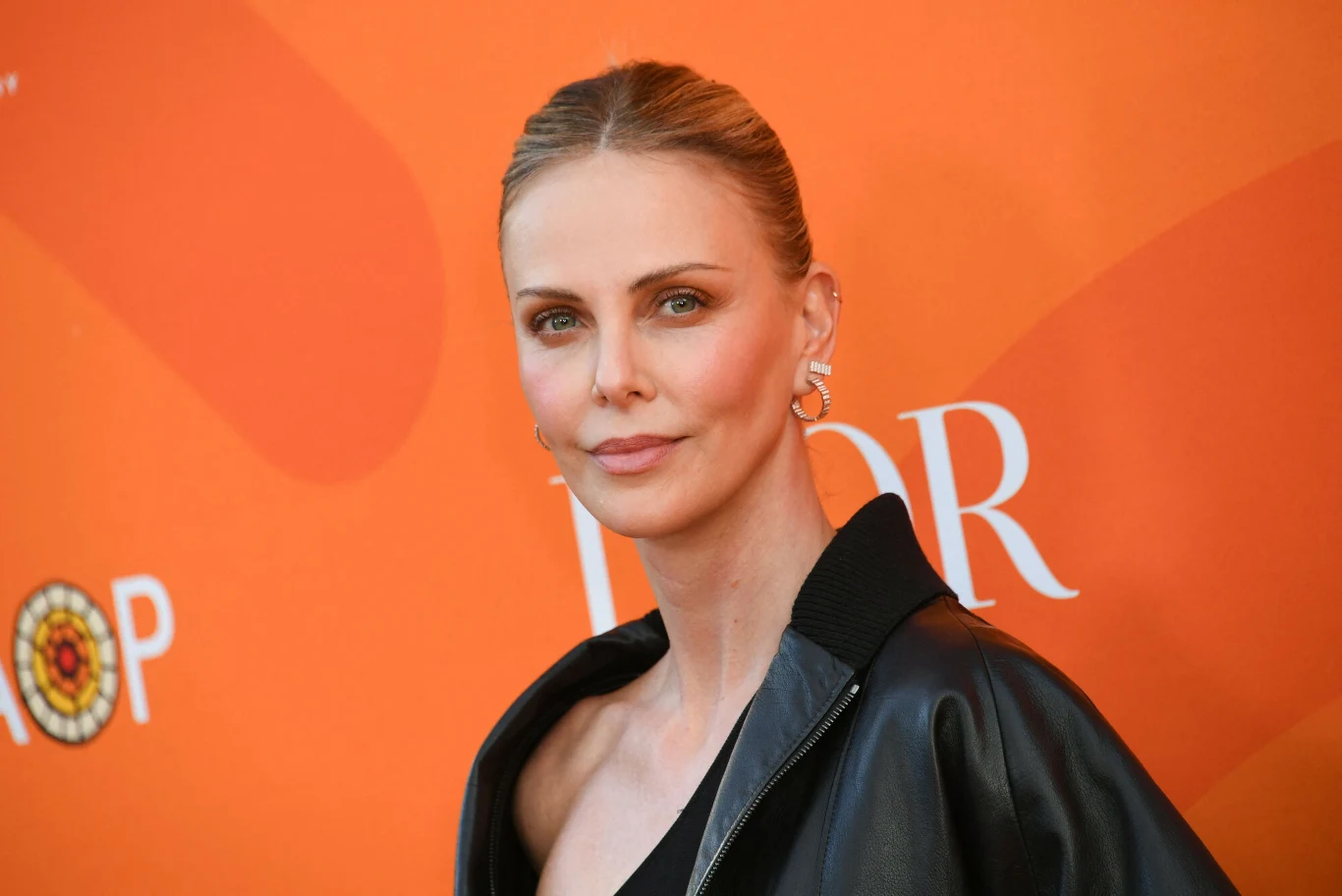 Charlize Theron zachwyca perfekcyjną sylwetką Charlize Theron z jasnymi spiętymi włosami, w czarnej kurtce i dużych kolczykach, na tle pomarańczowej ścianki z napisem