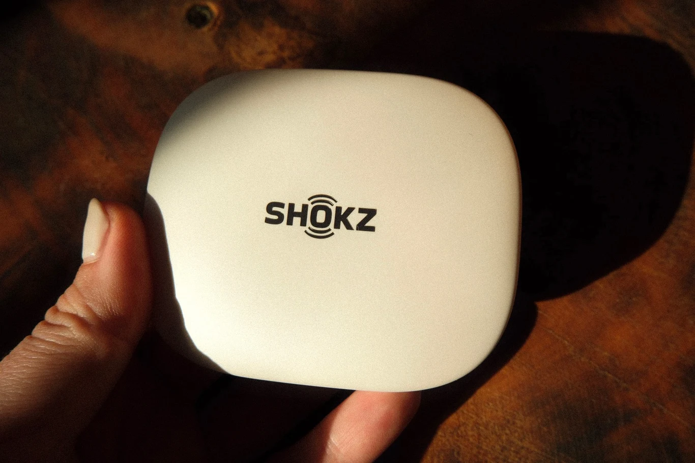 Etui ładujące Shokz OpenFit Pro Jasne, kompaktowe etui z logo SHOKZ trzymane w lewej dłoni, oświetlone ciepłym światłem.