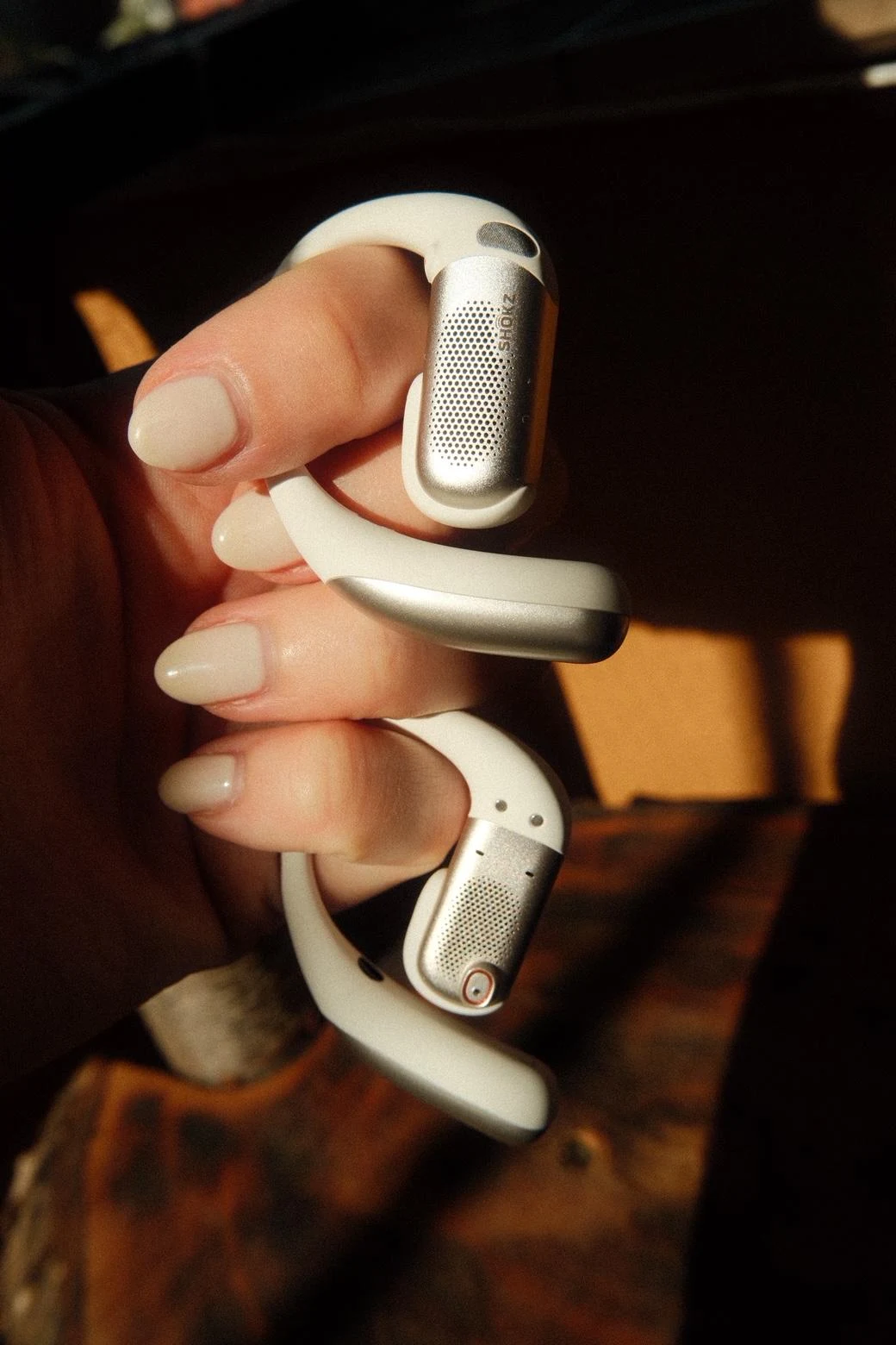 Shokz OpenFit Pro w wersji białej Dłoń trzymająca dwa bezprzewodowe zestawy słuchawkowe z mikrofonem w kolorze białym, o opływowym kształcie i metalicznych elementach.