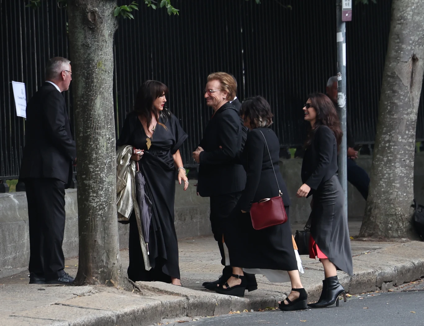 Bono przed katedrą św. Patryka w Dublinie przed rozpoczęciem uroczystości pogrzebowych Glena Hansarda Grupa elegancko ubranych osób rozmawia przy chodniku w cieniu drzew, dominują ciemne, stonowane kolory ubrań, jedna z kobiet trzyma czerwoną torebkę.