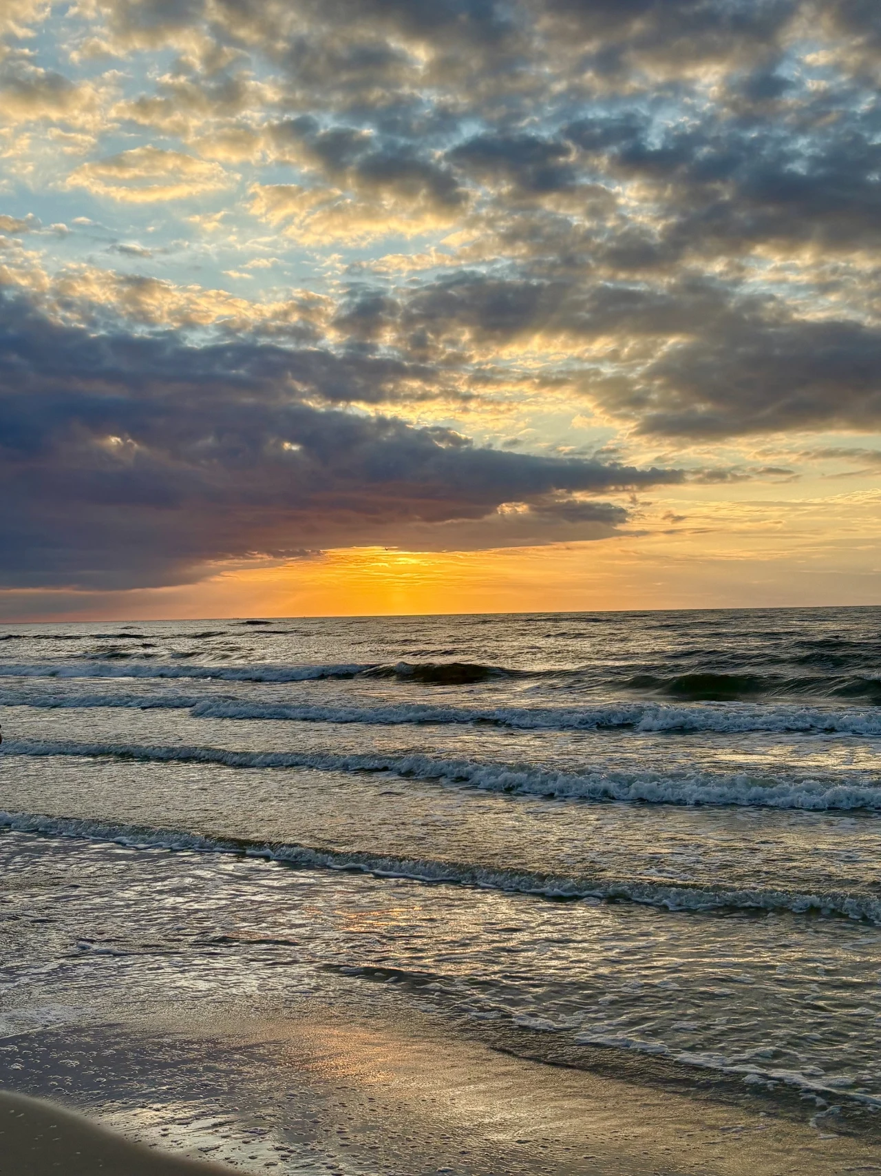 Zachodzące słońce rozświetla chmury nad wzburzonym morzem, a fale delikatnie rozbijają się o piaszczystą plażę.