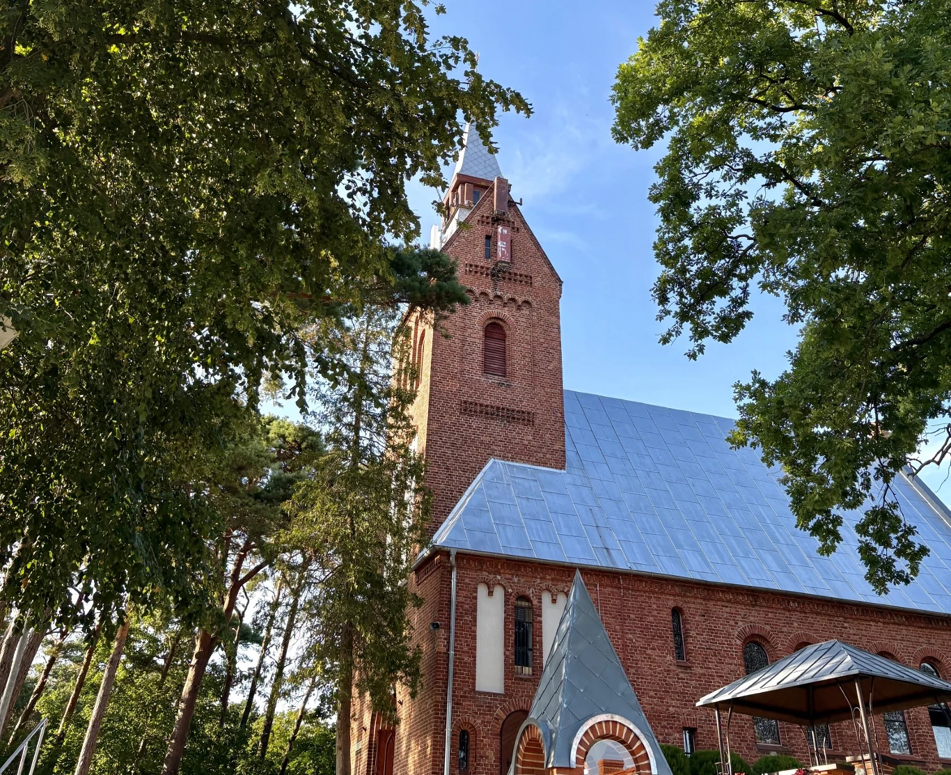 Ceglana wieża kościoła z wysokim dachem pokrytym blachą, otoczona wysokimi drzewami, na pierwszym planie widoczny fragment altany oraz niewielki spiczasty dach wejścia.