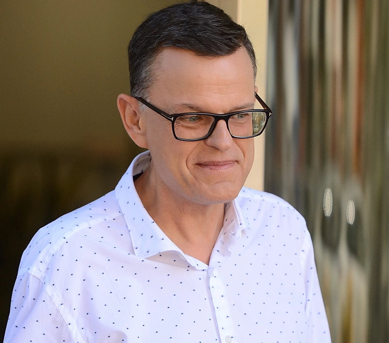 Andrzej Morozowski przed studiem TVN Mężczyzna w okularach, ubrany w jasną koszulę w drobne kropki, z krótkimi ciemnymi włosami, stoi w pobliżu przeszklonej ściany.