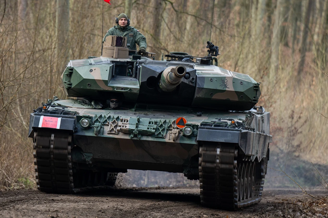 Czołg Leopard 2A5 z polskim żołnierzem w wieżyczce przemieszcza się leśną drogą, otoczony drzewami bez liści.