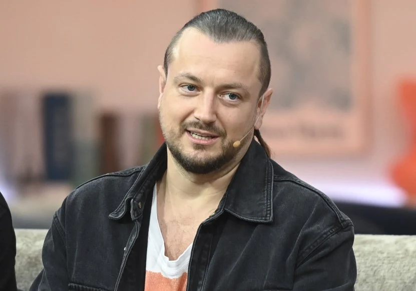 Mężczyzna z brodą i długimi, zaczesanymi do tyłu włosami, ubrany w czarną kurtkę jeansową, siedzi na kanapie z mikrofonem zamocowanym przy twarzy.