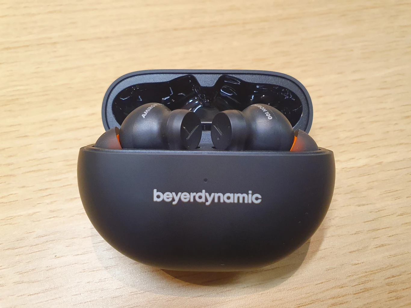 Bezprzewodowe słuchawki douszne marki beyerdynamic w czarnym etui ładującym, ustawione na jasnym, drewnianym blacie.