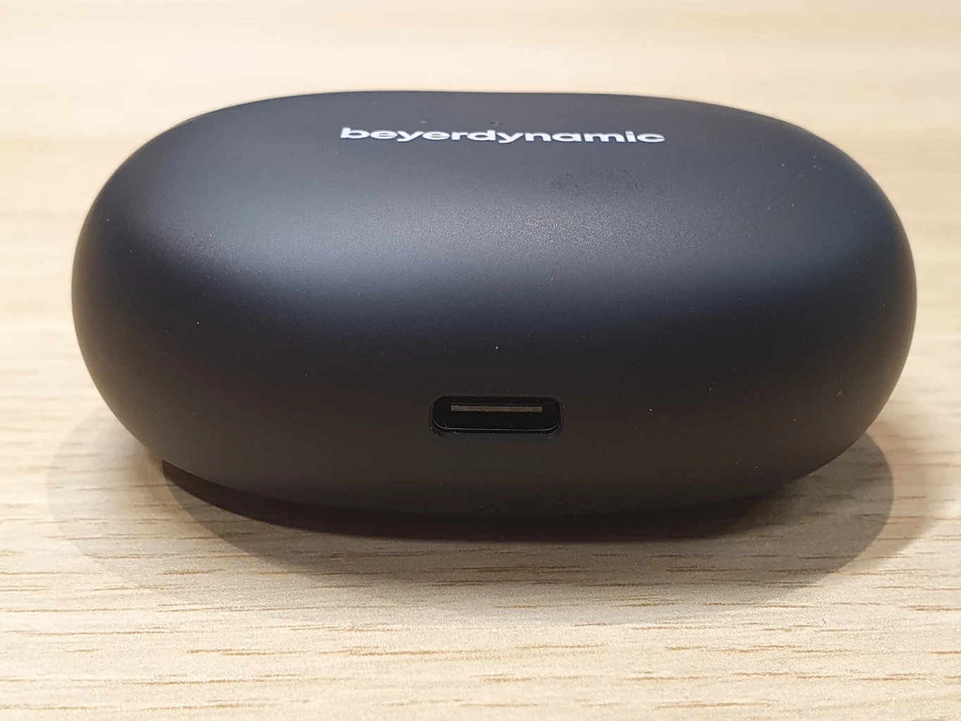Czarne etui ładujące do słuchawek bezprzewodowych Beyerdynamic z portem USB-C, ustawione na jasnym drewnianym blacie.