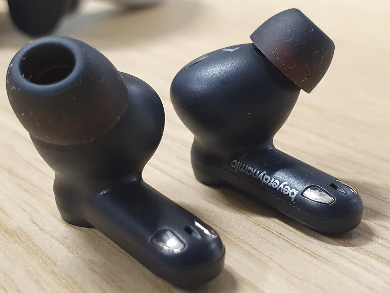 Dwie czarne bezprzewodowe słuchawki dokanałowe marki Beyerdynamic leżące na drewnianej powierzchni.