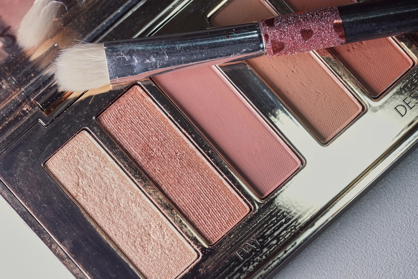 Paleta cieni do powiek w odcieniach beżu, różu i brązu oraz pędzel do makijażu z białym włosiem na tle zamkniętej kasetki.