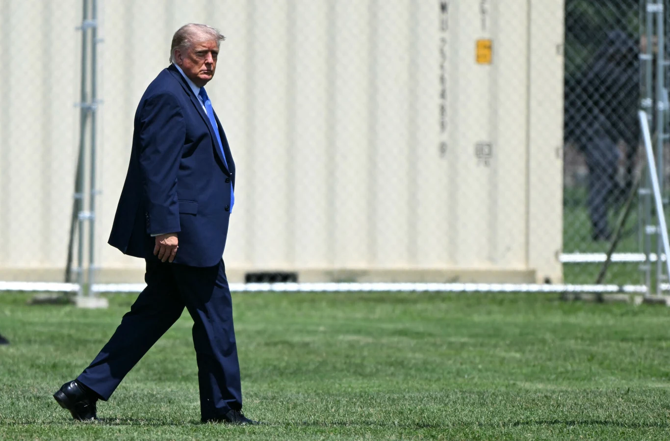 Donald Trump Donald Trump w ciemnym garniturze idzie po zielonej trawie obok jasnego kontenera i metalowego ogrodzenia.