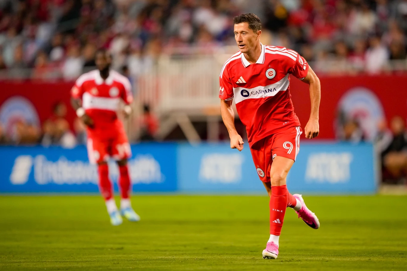 Robert Lewandowski podczas meczu Chicago Fire Robert Lewandowski w czerwonym stroju Chicago Fire z numerem 9 biegnie po boisku, obok inny zawodnik i widownia.