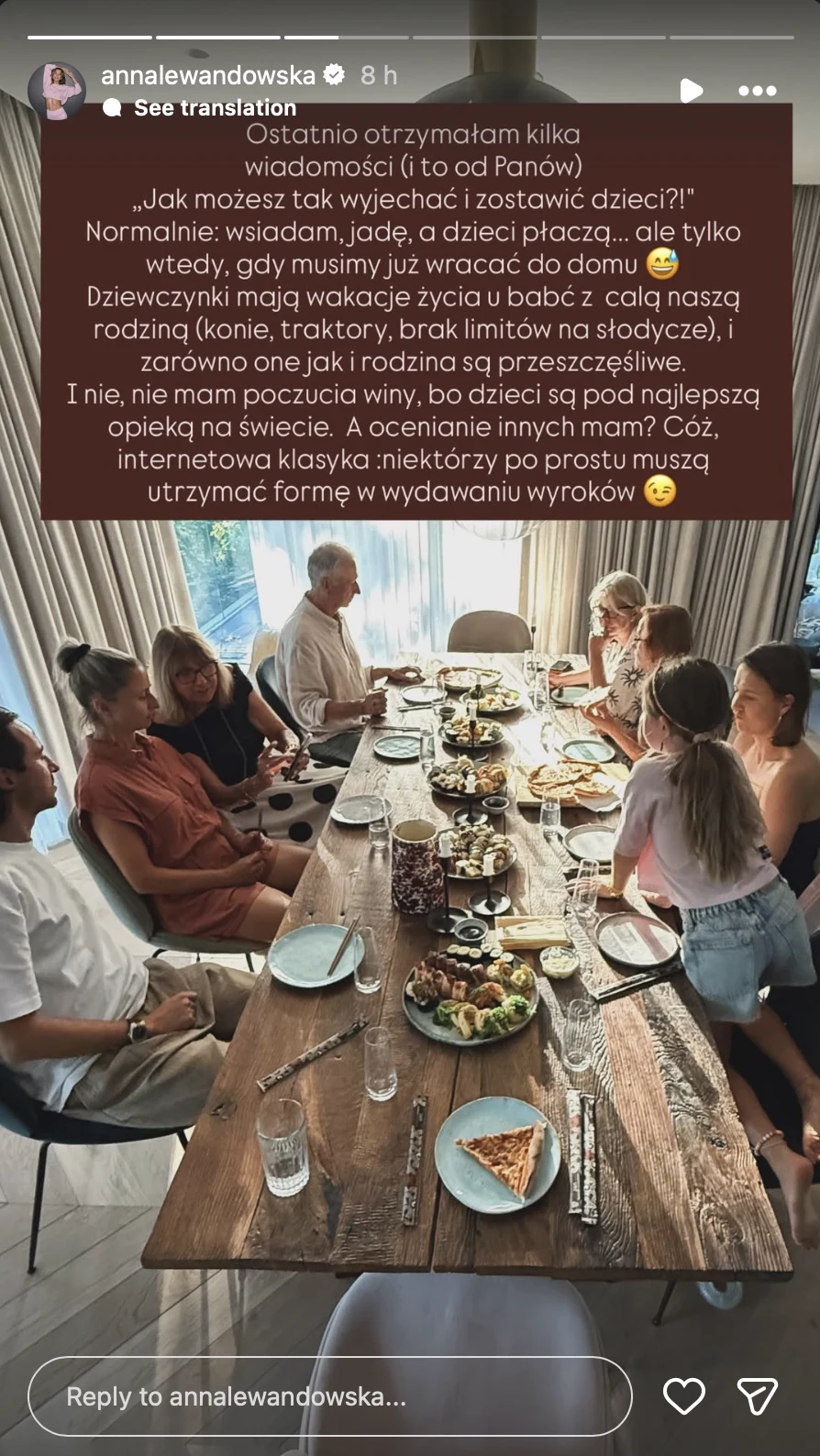 Grupa dorosłych i dzieci siedząca przy dużym, drewnianym stole, na którym rozstawione są liczne talerze z jedzeniem, napoje oraz owoce, w jasnym, przestronnym pomieszczeniu z dużym oknem i naturalnym światłem.
