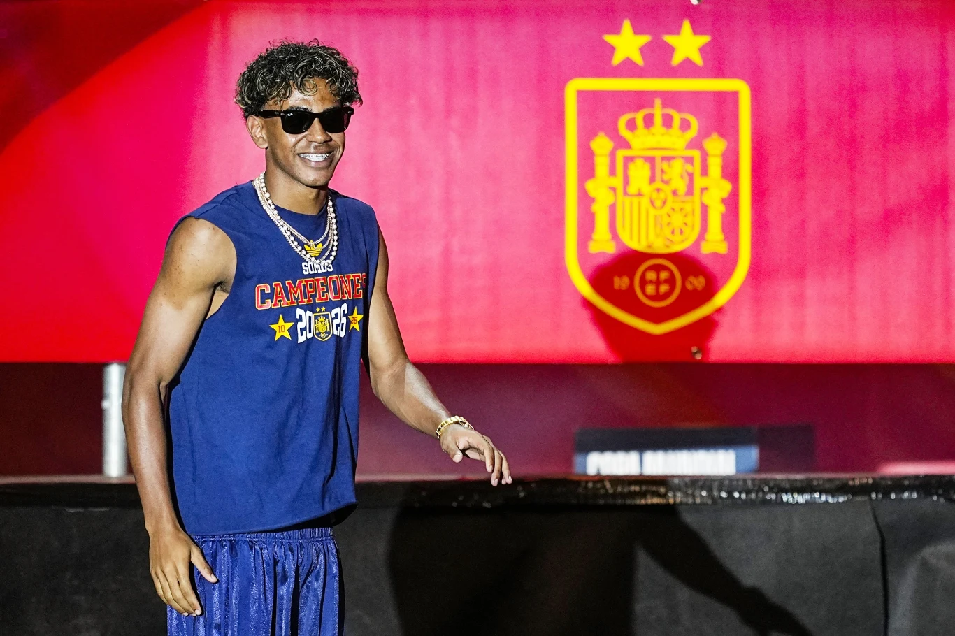 Lamine Yamal Mężczyzna w okularach przeciwsłonecznych i sportowej koszulce z napisem „Campeones” uśmiecha się, stojąc przed dużym godłem Hiszpanii z dwiema gwiazdkami.