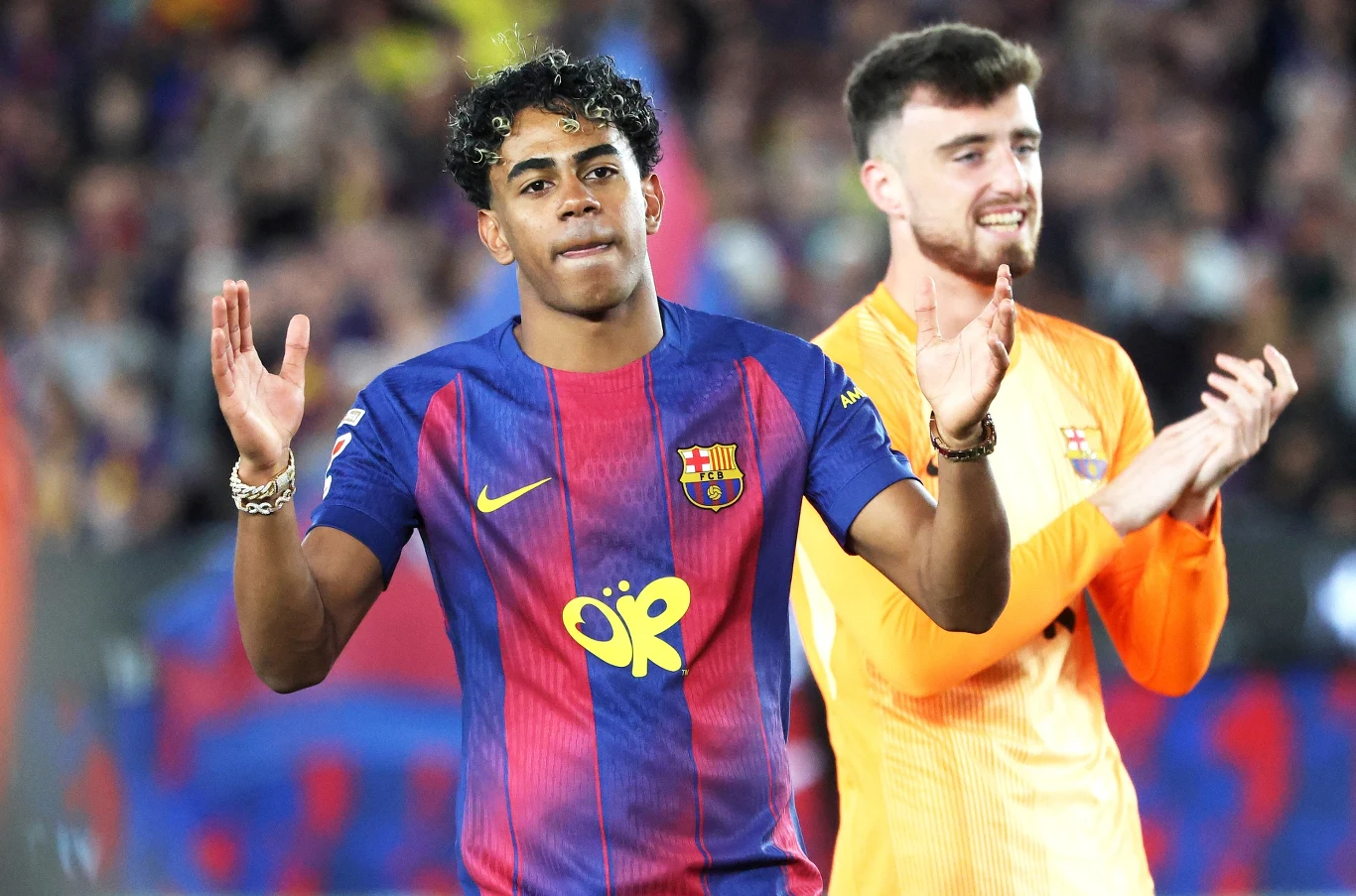 Lamine Yamal Zawodnik FC Barcelony w granatowo-bordowej koszulce z podniesionymi rękami obok bramkarza w żółtym stroju, obaj na tle rozmytego stadionu z kibicami.