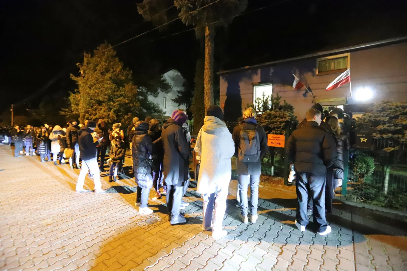 Grupa ludzi stojących wieczorem w długiej kolejce przed budynkiem ozdobionym polską flagą i oznaczeniem lokalu wyborczego.