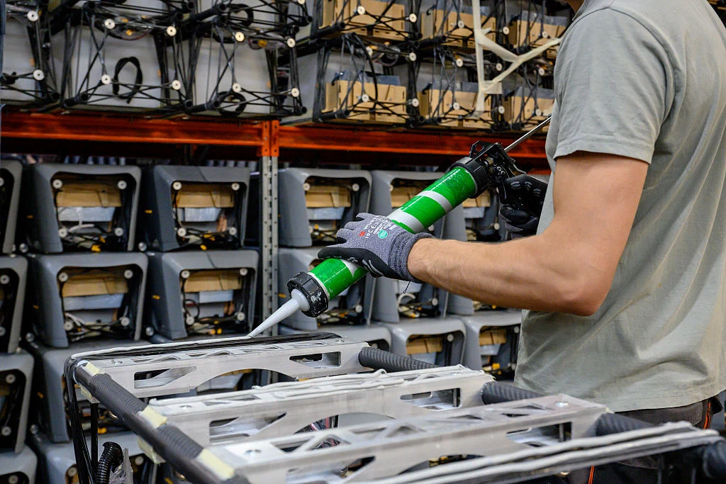 Pracownik w rękawicach nakłada uszczelniacz na metalową ramę, w tle widoczne są rzędy złożonych konstrukcji aluminiowych poukładanych na industrialnych regałach.