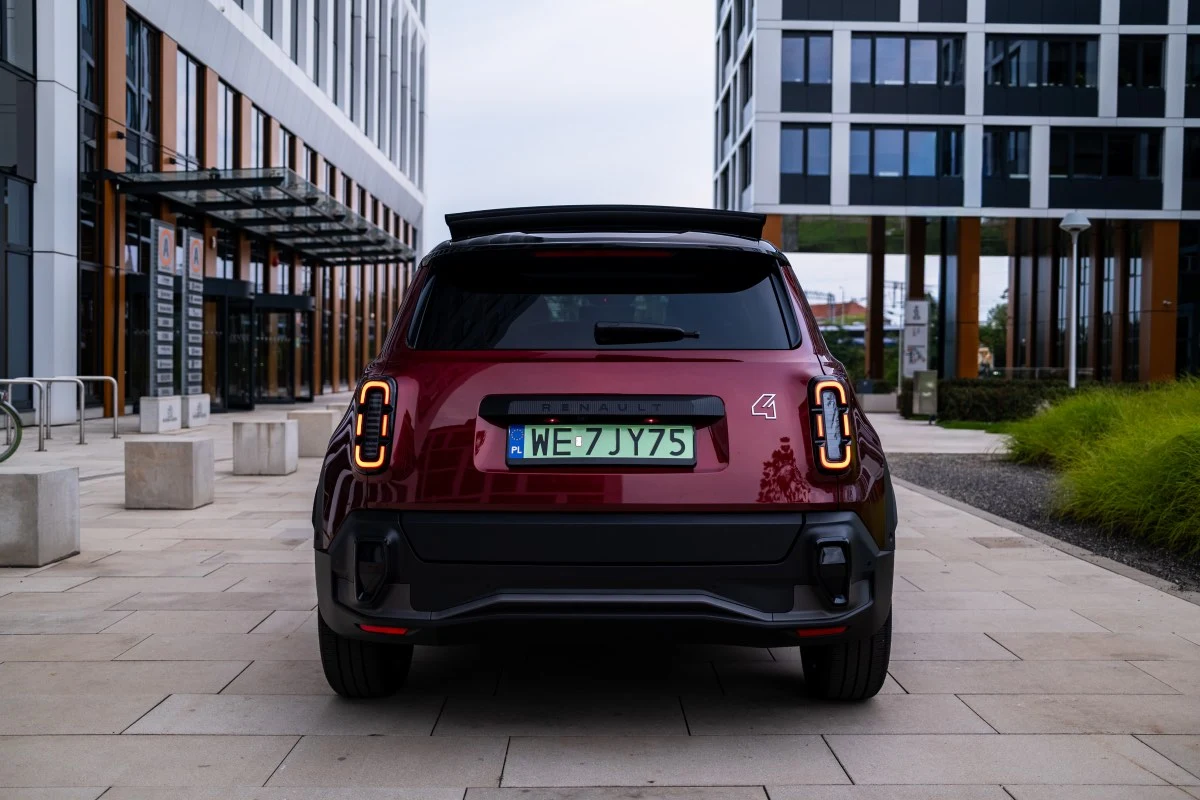 bordowy samochód elektryczny marki MINI z zielonymi tablicami rejestracyjnymi, widok od tyłu, zaparkowany między nowoczesnymi budynkami