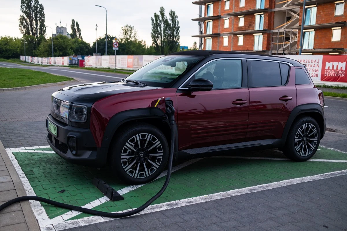Czerwony samochód elektryczny podłączony do stacji ładowania na wyznaczonym zielonym miejscu parkingowym w pobliżu budynku mieszkalnego w trakcie budowy.